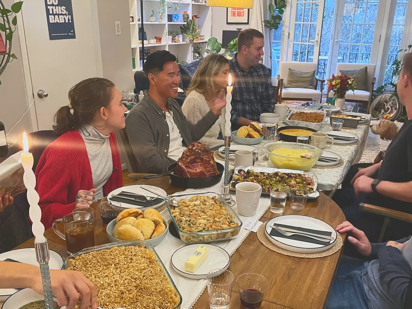 Friends passing dishes around a candlelit holiday table with glazed ham, stuffing and mac and cheese