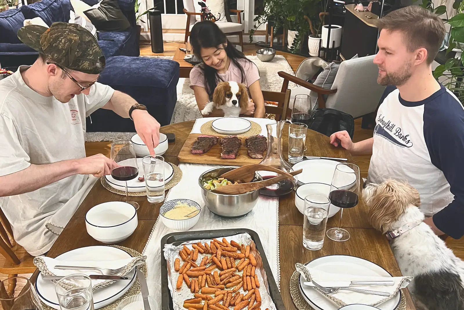 Friends sharing a dinner of sliced steak, roasted carrots and salad, with two dogs hoping for scraps
